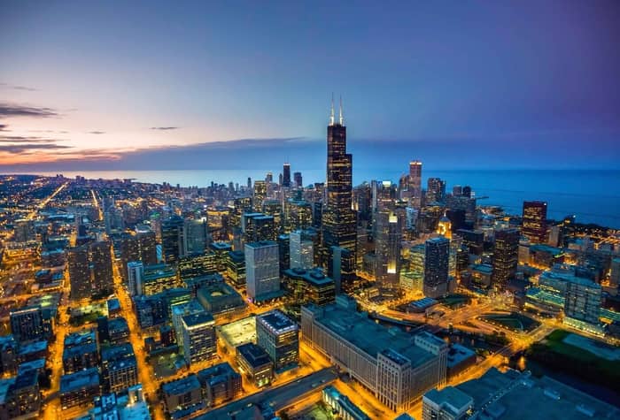 An aerial view of a cityscape at dusk, with lights illuminating the buildings and streets. The city is situated next to a body of water, possibly a lake or ocean.