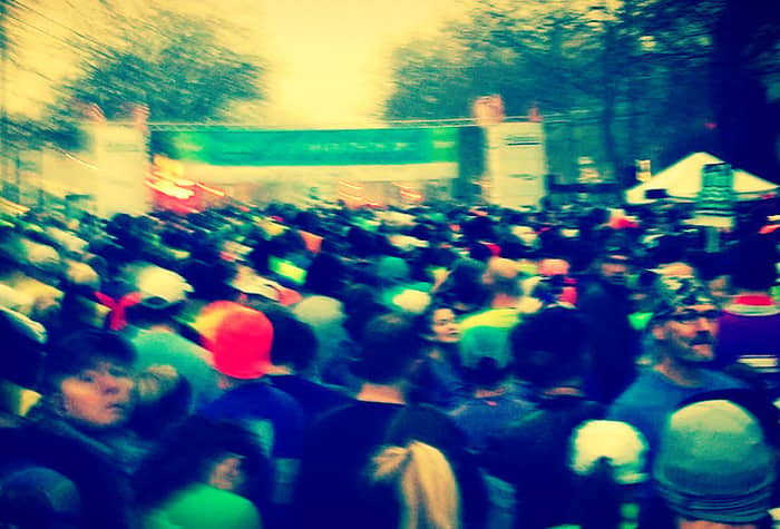 A blurry photo of a large crowd of people, likely at a race or marathon event, gathered near a banner and tents.
