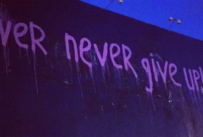 A photo of a billboard or wall with the phrase "Never Never Give Up!" painted on it in dripping purple paint. A figure appears to be painting on the wall in the bottom right corner.