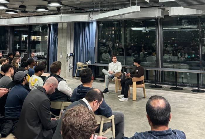 A photo of a group of people sitting in an indoor space, seemingly listening to two men who are seated in chairs at the front.