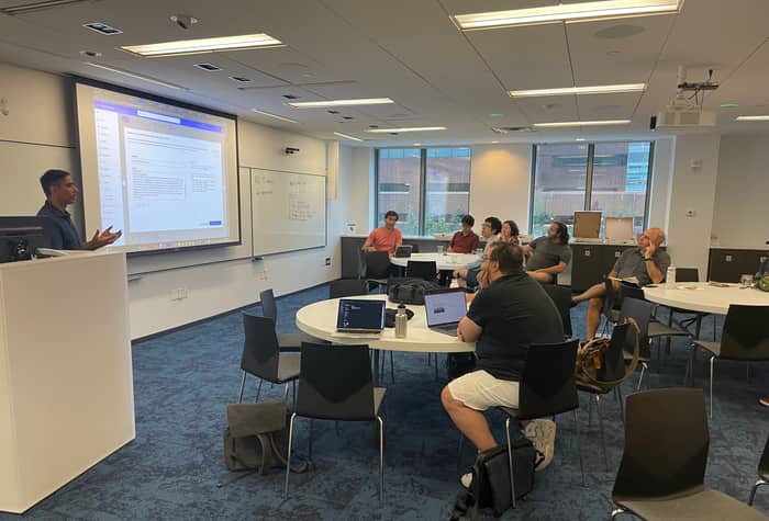 A photo of a presentation taking place in a conference room. A presenter stands at a podium, gesturing towards a projected screen, while an audience sits at round tables.