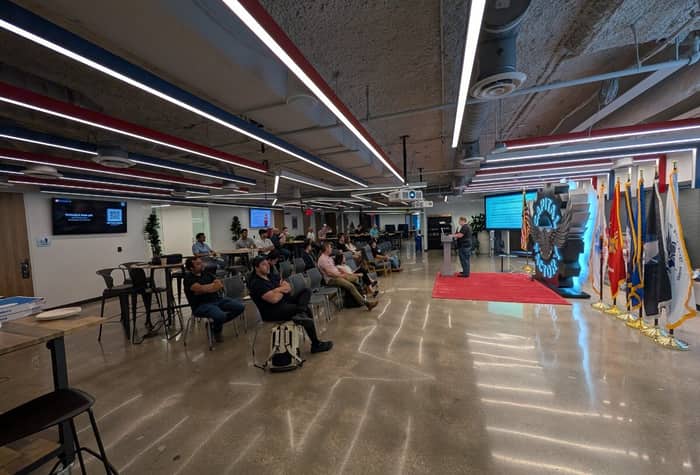 A man delivers a presentation to a seated audience in a modern, industrial-style event space featuring a large 'Capital Factory' logo and a row of military flags.. Text: CAPITAL FACTORY, UNITED STATES COAST GUARD