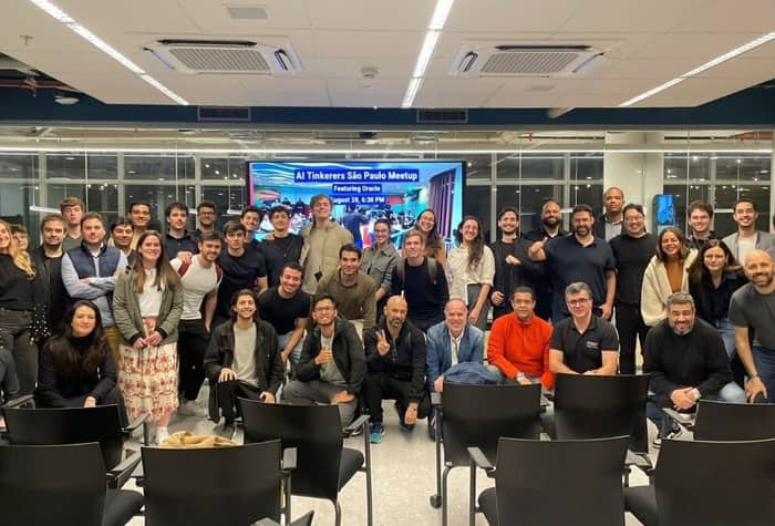 A group of approximately 40-50 people, appearing to be participants or attendees of an event, are posing for a photo indoors. A large screen in the background displays event details for an 'AI Tinkerers São Paulo Meetup'.. Text: AI Tinkerers São Paulo Meetup
Featuring Oracle
August 28, 6:30 PM