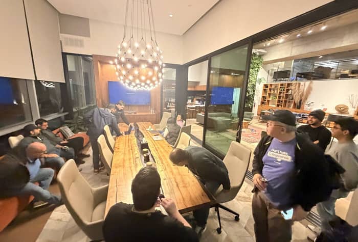 A candid photograph captures a group of approximately ten people gathered around a long, rustic wooden conference table in a modern, well-lit indoor meeting space.. Text: Don't hate, Regenerate.