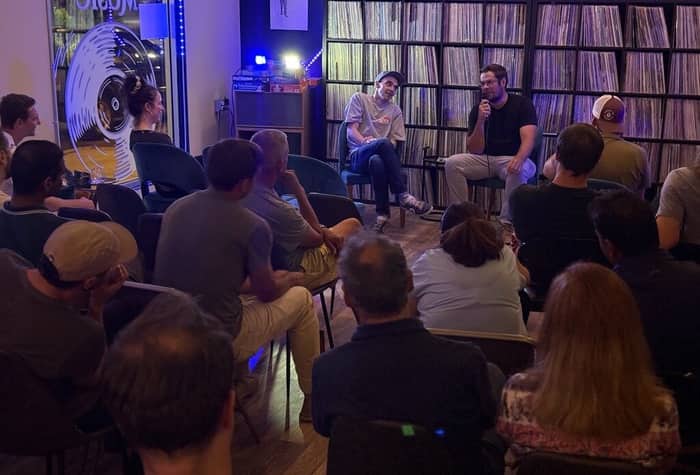 An indoor event showing two men speaking to a seated audience in front of shelves filled with vinyl records.. Text: MUSIC