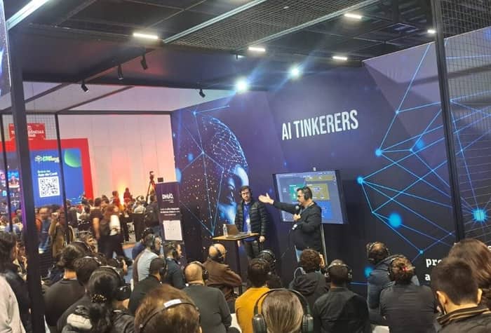 The image captures a technical presentation or workshop session taking place at a large indoor conference or exhibition, featuring a seated audience wearing headphones and two speakers standing in front of a dark blue backdrop labeled 'AI TINKERERS.' The backdrop is decorated with abstract blue network and node graphics.. Text: AI TINKERERS ORACLE DWG Entre na App do [unreadable] pro biens rs. Dr. [unreadable] [other unreadable text]