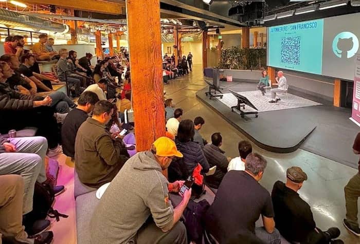A photo of an audience watching a presentation in a large, modern space. Two presenters are on a stage in front of a large screen displaying a QR code and a logo.. Text: #Auth0USERS - SAN FRANCISCO