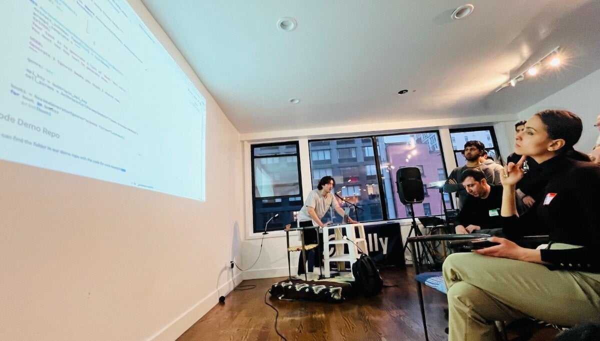 (Photo) A photo of a presentation taking place in a room with a projector screen displaying code and a speaker at a table facing an audience. Text: ode Demo Repo people | indoor | presentation, projector screen, audience, speaker | candid Note: The image is a real-life captured image of a presentation, depicting people and a setting. It's not a drawn or computer-generated image, thus classifying it as a photograph.