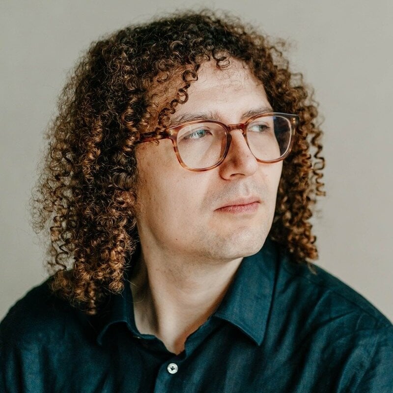(Photo) A portrait of a man with voluminous curly brown hair and glasses, looking thoughtfully to the side against a neutral background. person | indoor | curly hair, glasses, neutral background | portrait | Colors: #d8d1c6, #1a2a33, #7b5038 Note: The image is a clear, high-resolution photograph of a human subject, characteristic of a professional portrait or headshot.