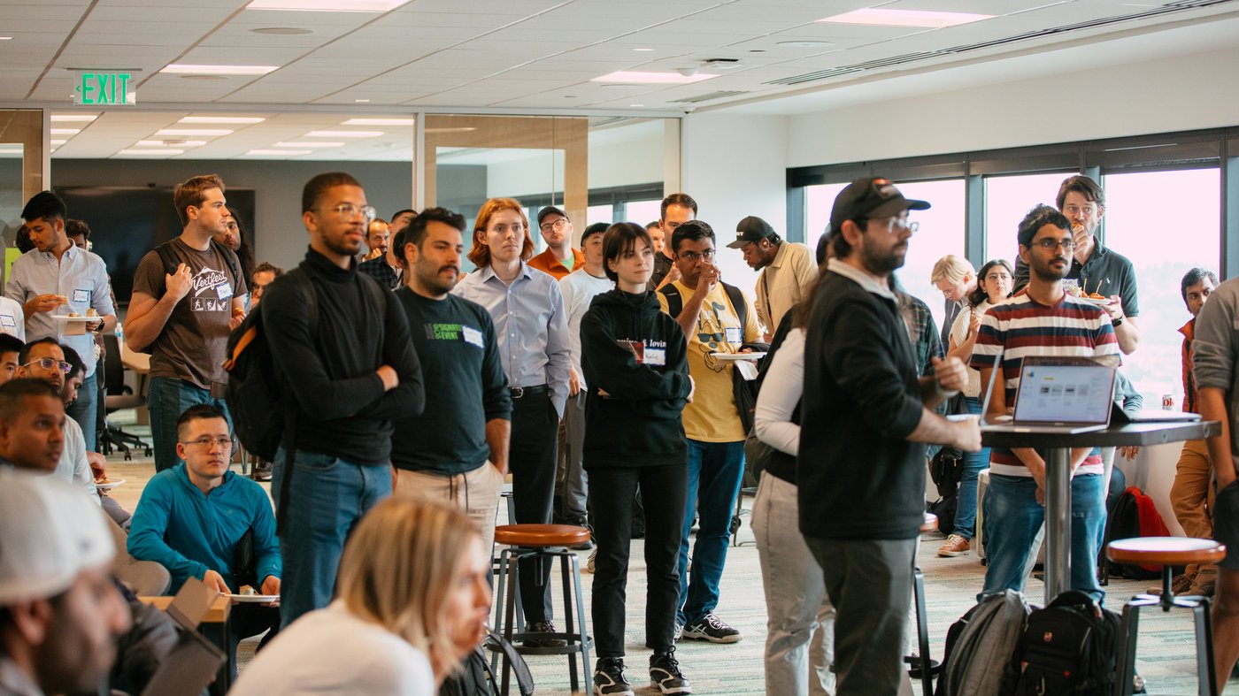 (Photo) A photograph of a group of people gathered in an office space, seemingly attending a presentation or meeting. Text: EXIT people | indoor | group of people, office setting, presentation/meeting | candid Note: The image is a real-life captured scene of people in an office environment, indicating a photograph. It depicts a spontaneous moment rather than a posed shot.