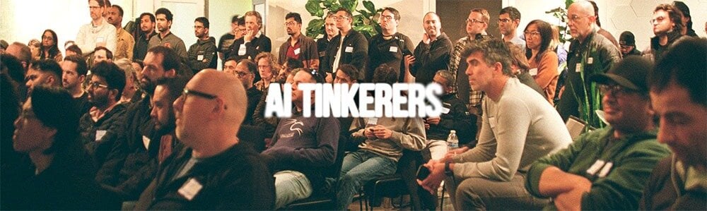 (Photo) This photo shows a large group of people seated and standing, likely attending a conference or meeting. The text 'AI TINKERERS' is superimposed over the image. Text: AI TINKERERS group of people | indoor | large crowd, superimposed text | candid | Colors: #4A4A4A, #A9A9A9, #FFFFFF Note: The image depicts a real-world scene with people, which is characteristic of a photograph. The text is added on top of the photo.