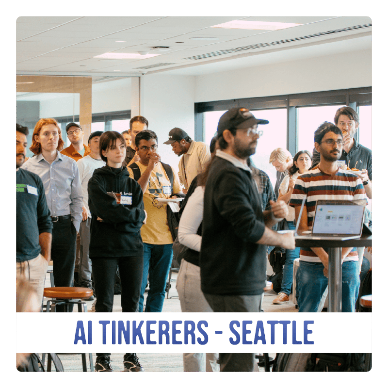 (Photo) A photograph of a group of people gathered in a large, modern room, seemingly at a conference or workshop. Text: AI TINKERERS - SEATTLE people | indoor | group of people, conference/workshop setting | candid Note: The image is a real-life captured scene of people in an indoor setting, making it a photograph. The text banner at the bottom provides context.