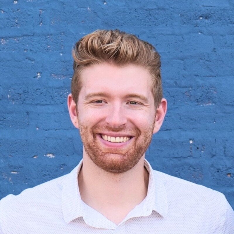 (Photo) A portrait photo of a smiling young man with light brown hair and a beard, posing in front of a blue brick wall. person | outdoor | smiling man, blue brick wall background, headshot portrait | portrait | Colors: #3b6da5, #f4f3f6, #b98a68 Note: The image is a real-world camera-captured portrait of a person, making 'photo' the most appropriate category.