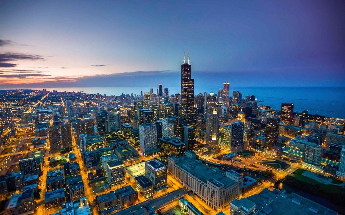 (Photo) An aerial view of a cityscape at dusk, with lights illuminating the buildings and streets. The city is situated next to a body of water, possibly a lake or ocean. cityscape | outdoor | skyline, body of water, city lights | aerial Note: The image is a real-life captured image of a city skyline, making it a photograph. It doesn't contain any text or design elements that would categorize it as a logo, banner, or illustration.