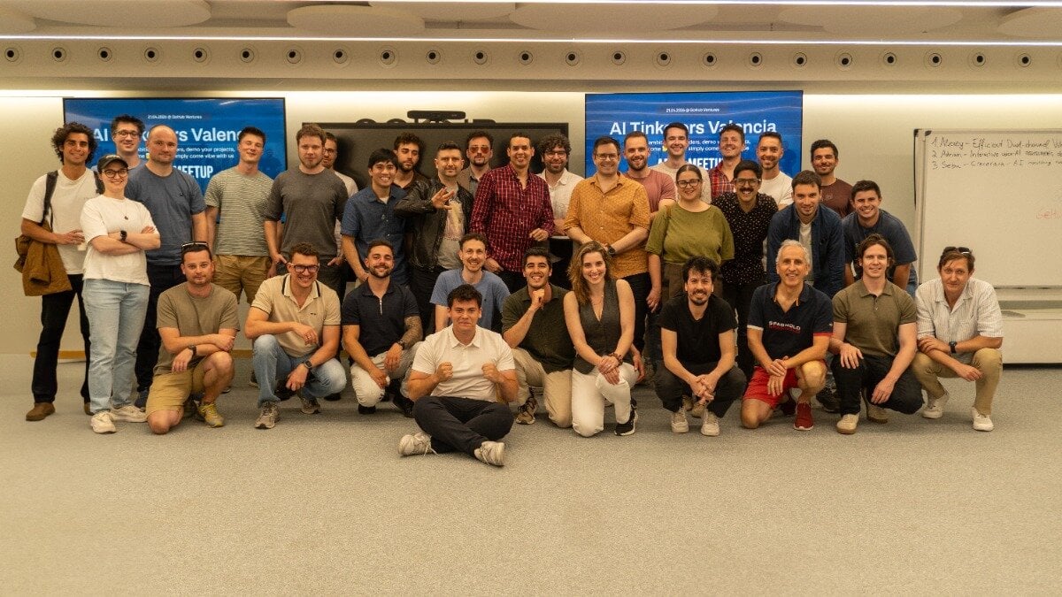 (Photo) A large group of people, mostly men, posing for a group photograph in an indoor event space with screens and a whiteboard in the background. Text: AI Tinkers Valencia MEETUP AI Tinkers Valencia AI MEETUP 1. [unreadable] - Efficient [unreadable] 2. [unreadable] - Interactive [unreadable] 3. [unreadable] - [unreadable] group of people | indoor | large group, digital screens displaying event info, whiteboard with handwritten notes | group portrait | Colors: #E6E1D3, #1B3F8B, #2D2D2D Note: The image is a standard group photograph capturing a gathering of individuals in a professional or social setting.
