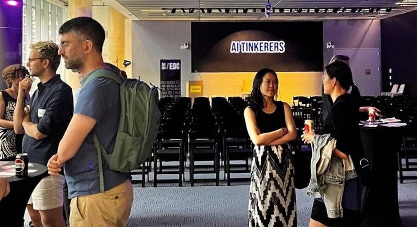 (Photo) A photo of people standing in a room with a banner that says 'AI TINKERERS'. Several people are standing around talking, and there are rows of chairs in the background. Text: AI TINKERERS /EDC people | indoor | group of people, banner, chairs | candid Note: This is a photograph of a real-life scene captured with a camera. It depicts people in a room, indicating a candid or event-style shot.
