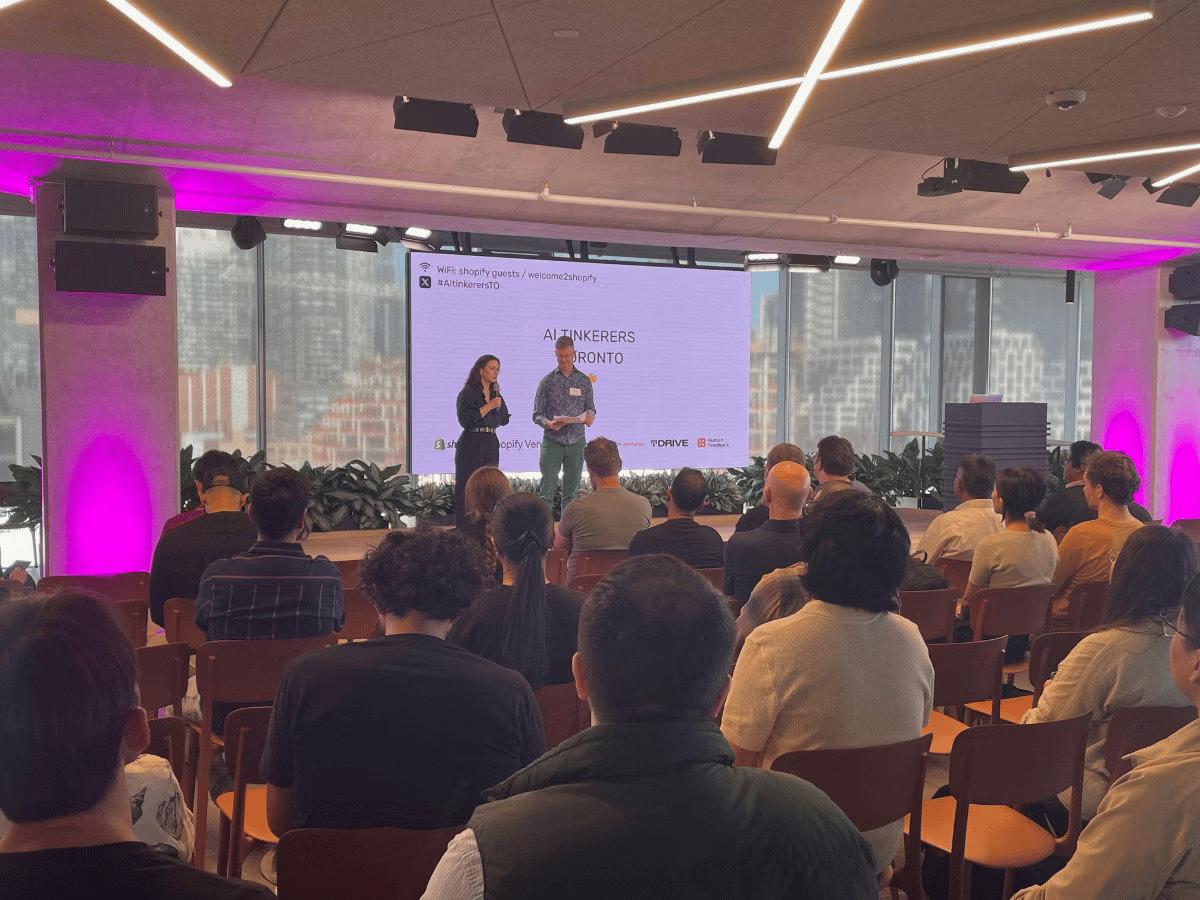 (Photo) A photograph capturing an indoor event, likely a tech conference or presentation, where two speakers are on a stage in front of a large screen displaying the title 'AI TINKERERS TORONTO.' A large audience is seated facing the stage, viewed from behind, in a modern venue with dramatic magenta lighting. Text: WiFi: shopify guests / welcome2shopify #AiTinkersTO AI TINKERERS TORONTO shopify Ventures People attending a presentation/conference | Indoor, modern event space with large windows overlooking a city skyline | Two speakers on stage, large projection screen, audience viewed from the back, dramatic magenta accent lighting. | Candid/event photography, wide shot | Colors: #FF00FF, #000000, #FFFFFF, #A0522D Note: The image is a direct capture of a real-world event, specifically a presentation or conference, showing the venue, speakers, and audience.