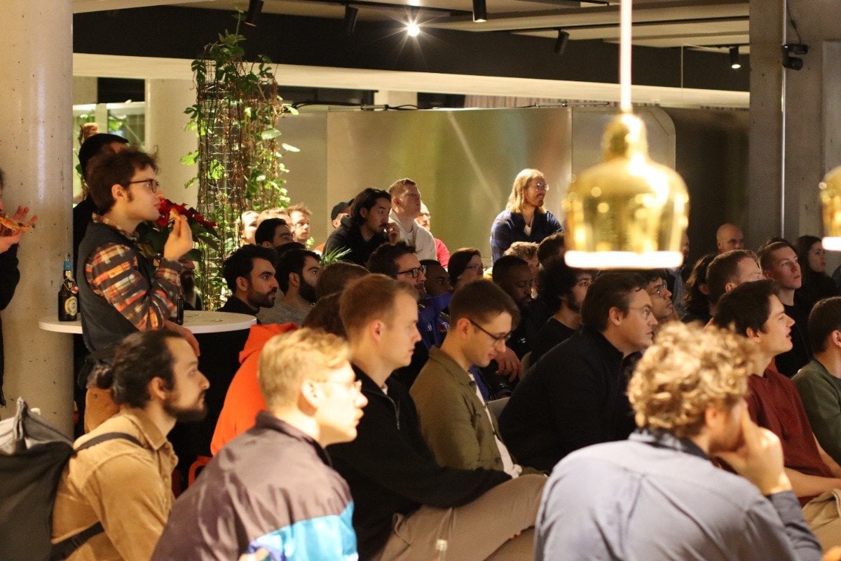 (Photo) A photo of a group of people sitting and standing in an indoor setting, appearing to watch a presentation or performance. people | indoor | group of people, casual setting, presentation/performance implied | candid Note: The image is a real-life captured moment of a group of people in a casual indoor environment, indicative of a photographic snapshot.