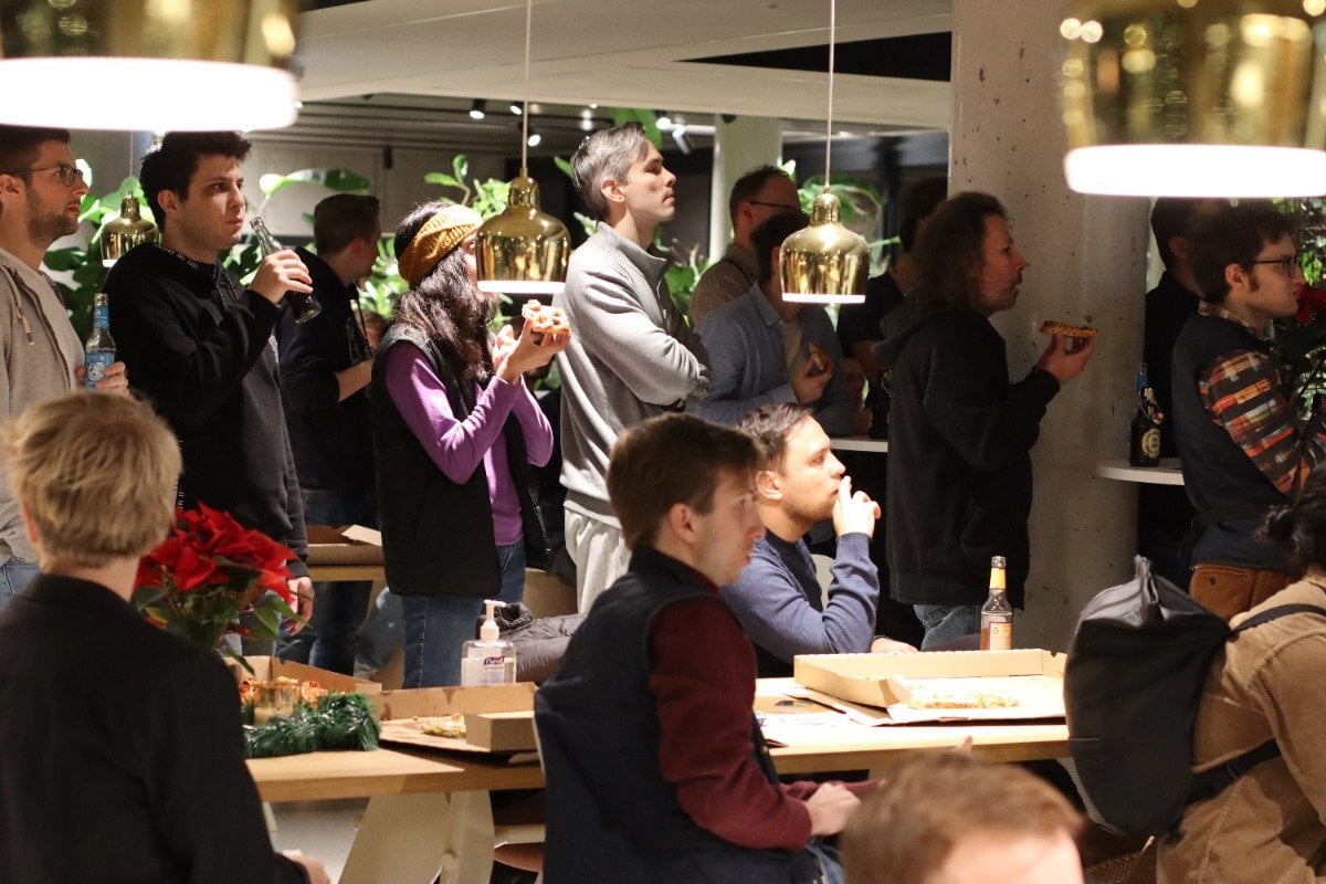 (Photo) A group of people are gathered in a brightly lit space, eating pizza and socializing. They are standing and sitting around tables. people | indoor | group of people, food, social gathering | candid Note: The image is a candid photograph of a real-life scene, capturing a moment in time.