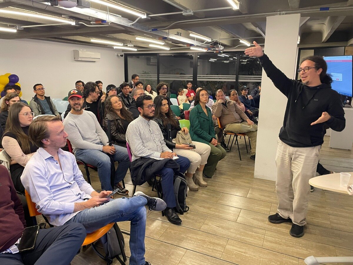 (Photo) A person stands and gestures while addressing a seated audience in a room. people | indoor | presentation, audience | candid Note: The image is a real-life captured moment of a presentation or talk, depicting people in a room.  This classifies it as a photograph.