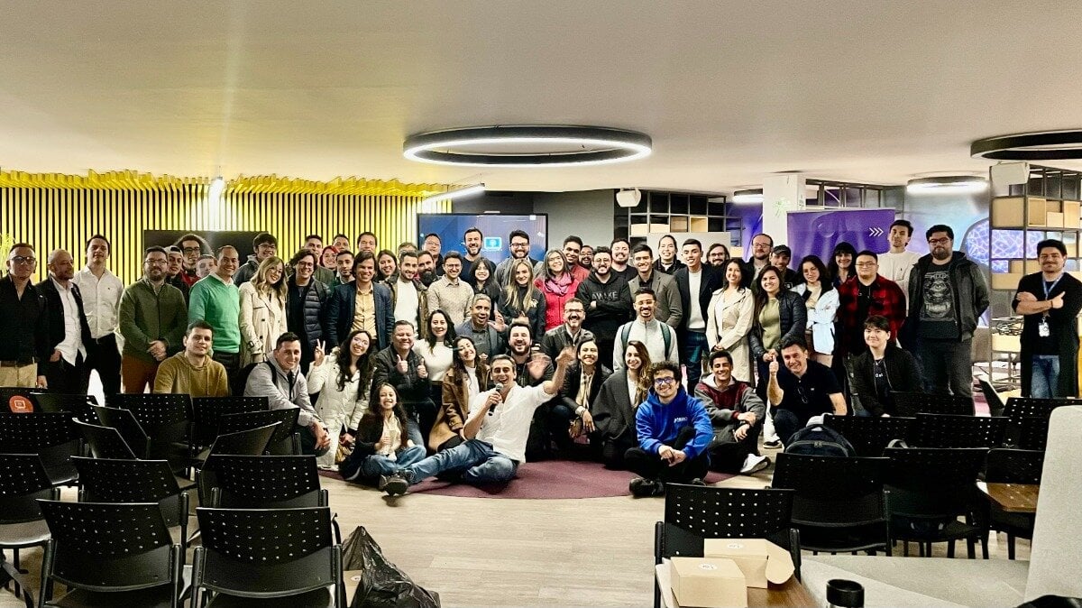 (Photo) A large group of people is posing for a photo in a modern office or event space.  Some are standing, some are sitting on the floor, and empty chairs are visible in the foreground. people | indoor | large group, office setting | group photo Note: This is a photograph capturing a group of people in a specific setting.  It's not a logo, illustration, banner, or handwriting.