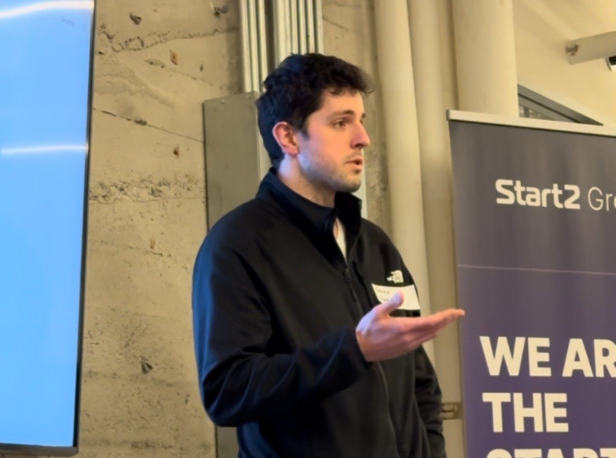 (Photo) A man is giving a presentation, gesturing with his hand. He is standing in front of a blue screen and a purple banner. Text: Start2 Gr WE ARE THE person | indoor | presentation, banner | candid | Colors: #420078, #007AFF, #231F20 Note: The image is a real-life captured moment of a person presenting, making it a photograph. It's not a staged studio shot, but rather an in-situ capture of an event.
