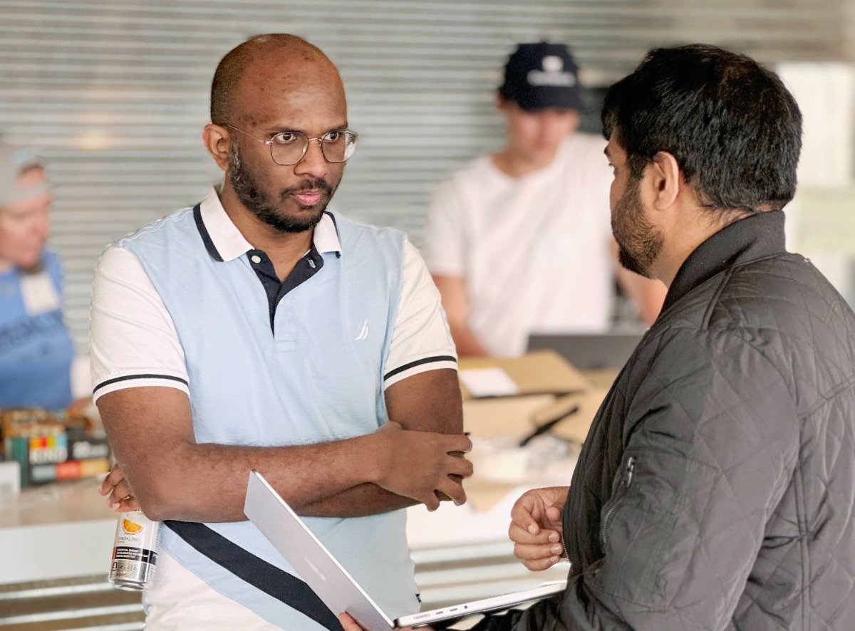 (Photo) Two men are having a conversation, one holding a laptop.  A third person is visible in the background. people | indoor | multiple people, conversation | candid Note: The image is a real-life captured image of people in a seemingly candid moment, indicative of a photograph.