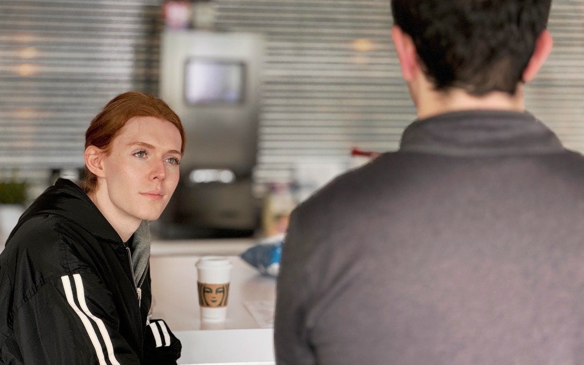 (Photo) A photograph of two people, one with red hair and the other with dark hair, seemingly in conversation at a counter or table. person | indoor | two people, casual conversation | candid Note: The image is a real-life captured moment, depicting people in a casual setting, which makes it a photograph.