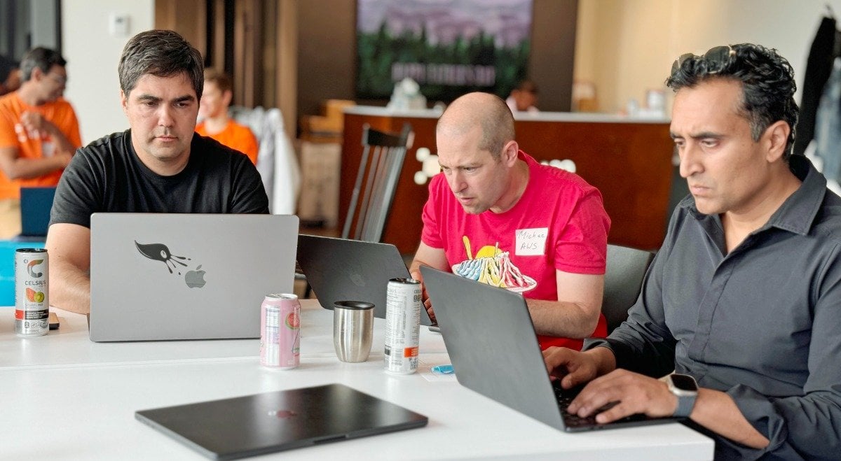 (Photo) Three men are sitting at a table, each focused on their laptops. They appear to be in a casual work or collaborative setting. Text: Michael AUS people | indoor | multiple people, laptops, casual setting | candid Note: The image is a real-life captured scene of people, clearly a photograph, not a digitally created image or other type.