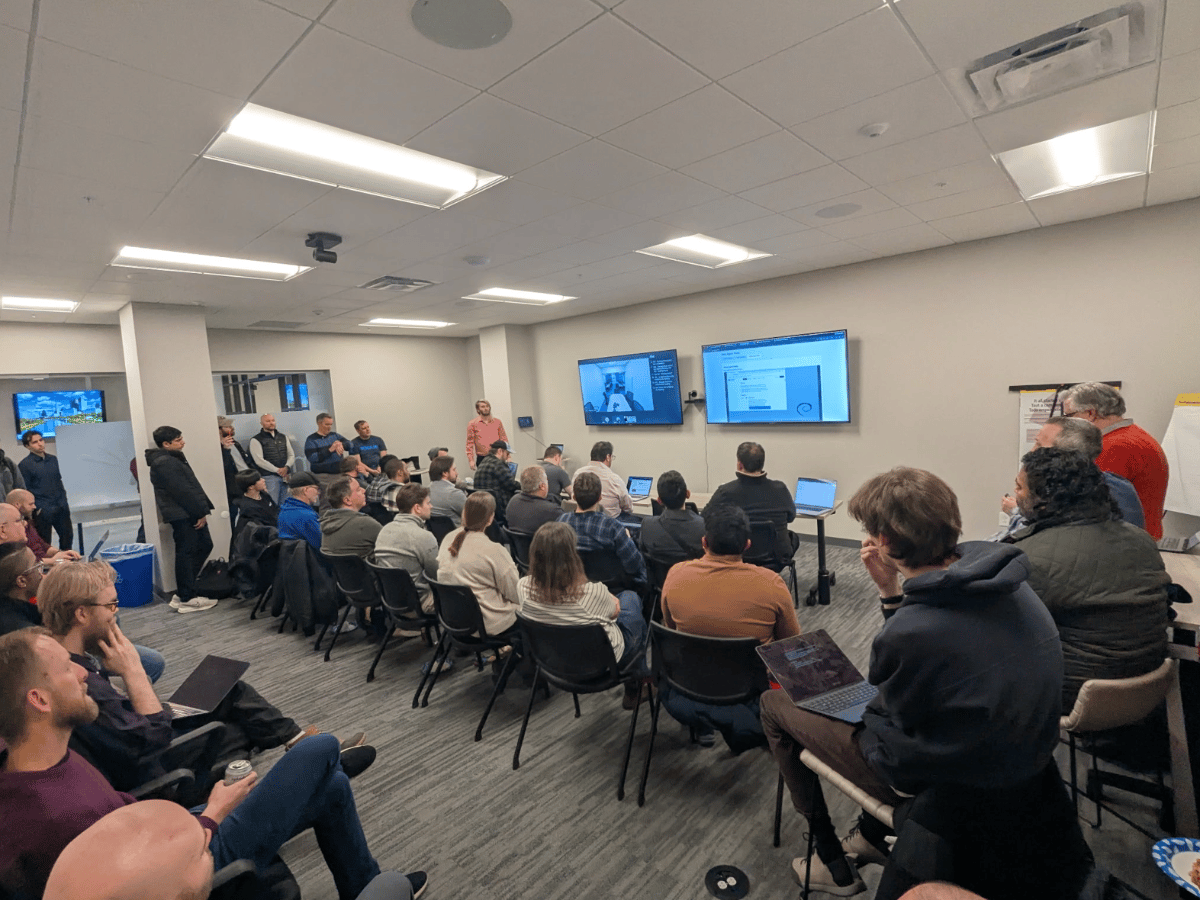 (Photo) A group of people are gathered in a conference room for a presentation, with two large screens displaying technical content and a Debian logo. people in a meeting or presentation | indoor office or conference room | multiple attendees, presentation screens, Debian logo, laptops | candid, wide-angle | Colors: #E0E0E0, #3C3C3C, #8B0000 Note: This is a real-life photograph capturing a group of people in an indoor setting during an event.
