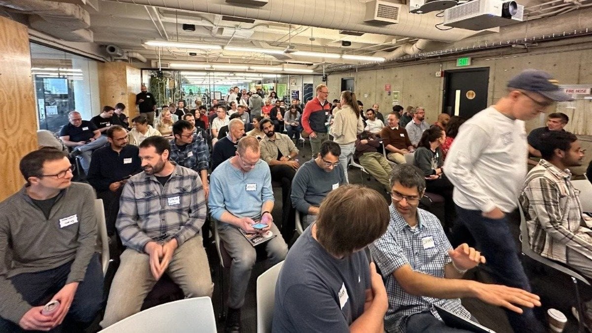 (Photo) A photo of a large group of people sitting and standing in a room, many looking at their phones or talking to each other. people | indoor | large group, some looking at phones | candid Note: The image is a real-life captured image of people in a room, indicative of a photograph. It's not a drawing, logo, or banner.
