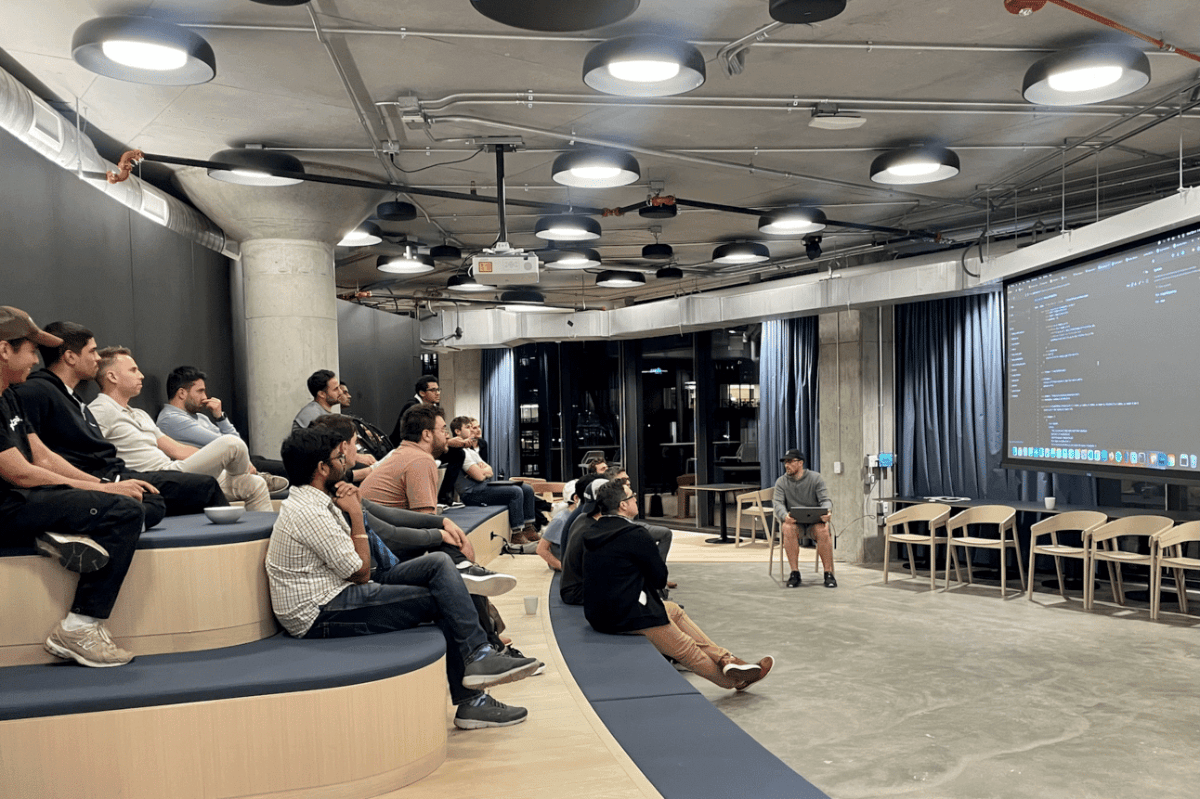 (Photo) A group of people are sitting in a modern office space, seemingly attending a presentation or training session.  One person is sitting on a chair with a laptop, possibly the presenter, while the others are seated on tiered, curved benches. people | indoor | group of people, presentation setting, modern office | candid Note: The image is a real-life captured scene of people in an office setting, indicative of a photograph. It's not a drawing, logo, or banner.
