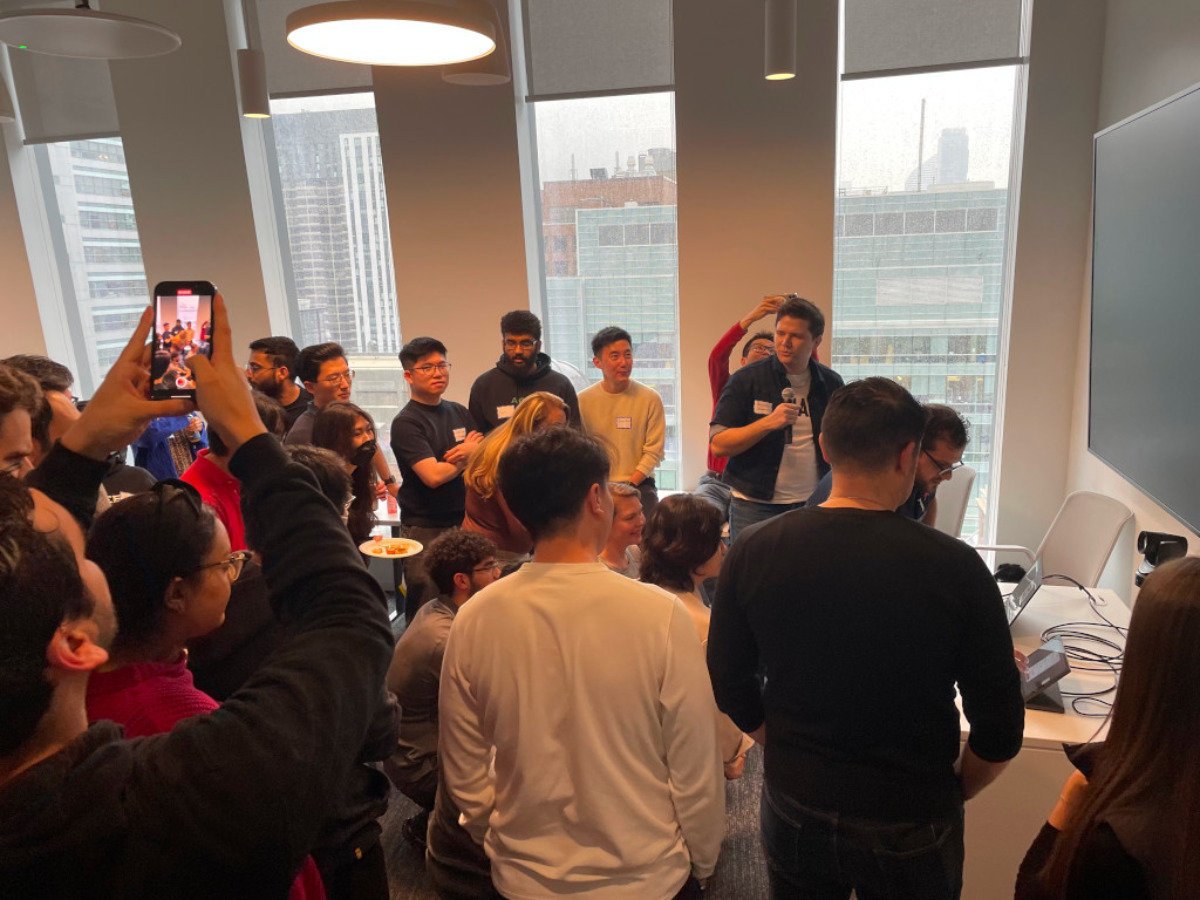 (Photo) A group of people are gathered in a room with large windows, some are listening to a speaker, and others are taking photos. people | indoor | group of people, presentation, taking photos | candid Note: The image is a real-life captured moment of a gathering, depicting people in a candid setting. This makes it a photograph.