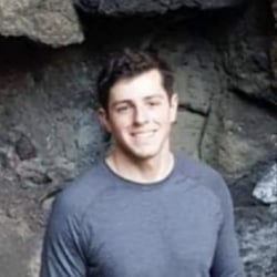 (Photo) A young man with short brown hair and a grey shirt smiles at the camera in front of a rocky background. person | outdoor | rocky background | portrait Note: The image is a real-life captured image of a person in a specific environment, making it a photograph.