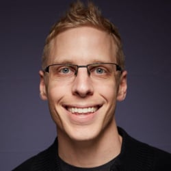 (Photo) A headshot photo of a smiling man with glasses and light brown hair. person | indoor | glasses, smile | portrait Note: The image is a real-life captured image of a person, making it a photograph. It is a headshot style portrait.