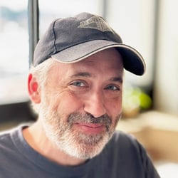 (Photo) A man with a grey beard and a baseball cap smiles at the camera. Text: REAT DIV person | indoor | beard, baseball cap | portrait Note: The image is a real-life captured image of a person, making it a photograph. It is not a drawing, logo, or other manufactured image type.