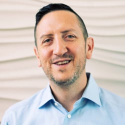 (Photo) A headshot of a man smiling at the camera. He is wearing a light blue, collared shirt and has short, dark hair. person | indoor | headshot, smiling | portrait | Colors: #ADD8E6, #E9E6DC Note: This is a real-life image of a person, captured by a camera, hence categorized as a photo.
