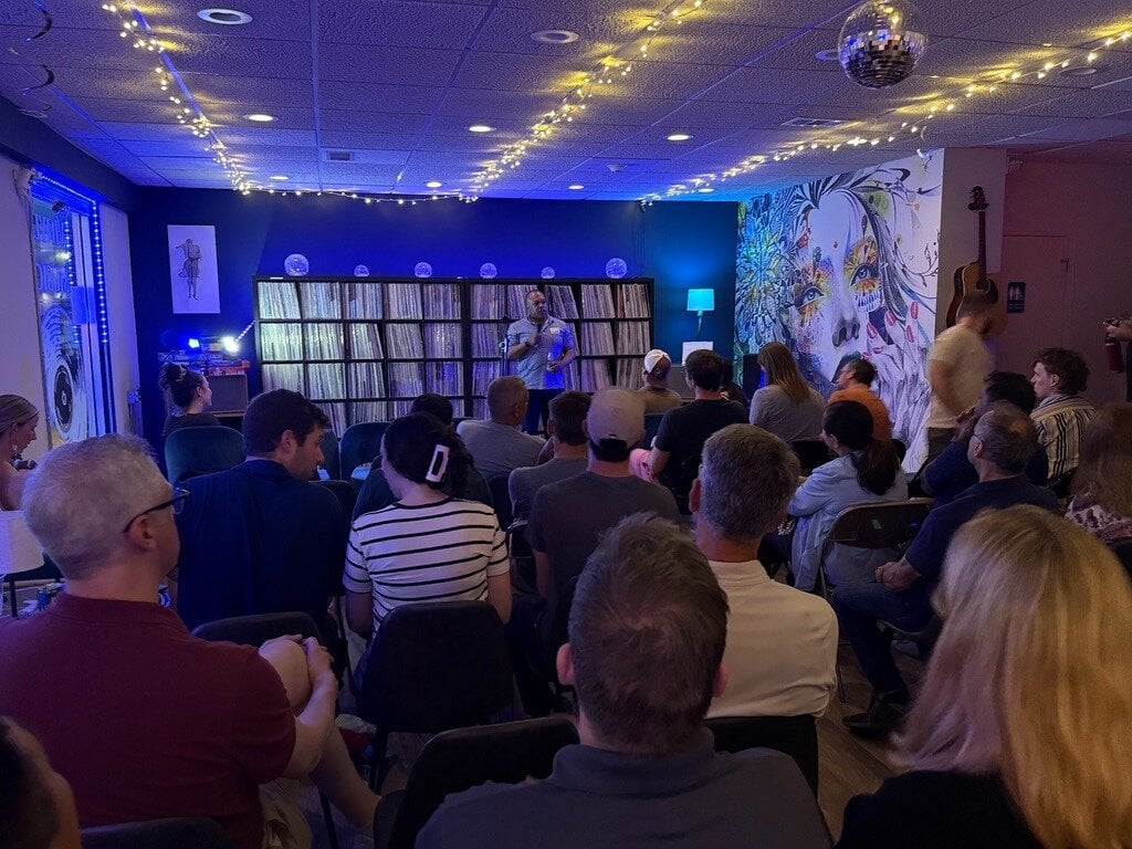 (Photo) A photo of an indoor presentation or gathering where a speaker is addressing a seated audience in a room with blue ambient lighting, string lights, and a large mural. presentation and audience | indoor | seated audience, speaker standing at the front, vinyl record shelves in the background, colorful wall mural, blue ambient lighting, string lights | candid | Colors: #121435, #3c4ca6, #e5d39c, #1e1e24 Note: This is a real-life photograph capturing a live event with a speaker and an audience in an indoor setting.
