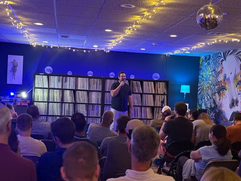 (Photo) A man stands on a stage holding a microphone, performing or speaking in front of a seated audience. The background features large shelves filled with vinyl records, illuminated by blue lighting, string lights, and a disco ball. stand-up comedy or speaking performance | indoor | audience, vinyl record shelves, blue ambient lighting, string lights, disco ball | candid | Colors: #1c244c, #0d111a, #a4b5d4, #e5e9f0 Note: This is a real-life photograph capturing a live indoor performance or event.