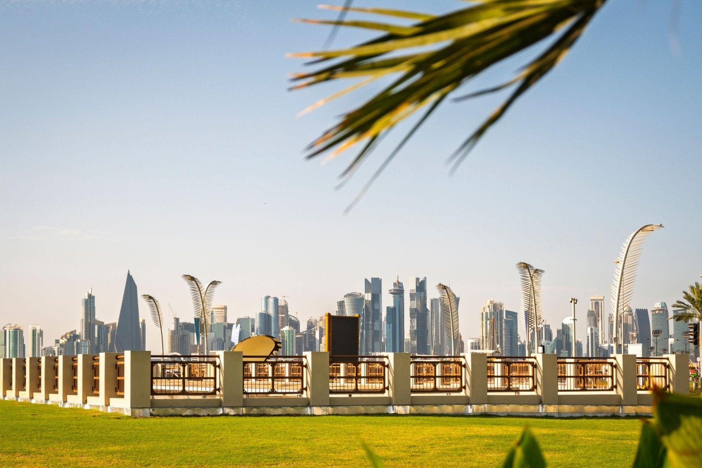 (Photo) A scenic view of a modern city skyline with skyscrapers, captured from a grassy park with a decorative fence and framed by palm leaves in the foreground. cityscape | outdoor | modern skyscrapers, grassy lawn, decorative fence, palm fronds framing the top | landscape photography | Colors: #9cb7d4, #8ba63c, #e5dec9, #4a5d6e Note: The image is a real-world photograph capturing a landscape and architectural cityscape scene.