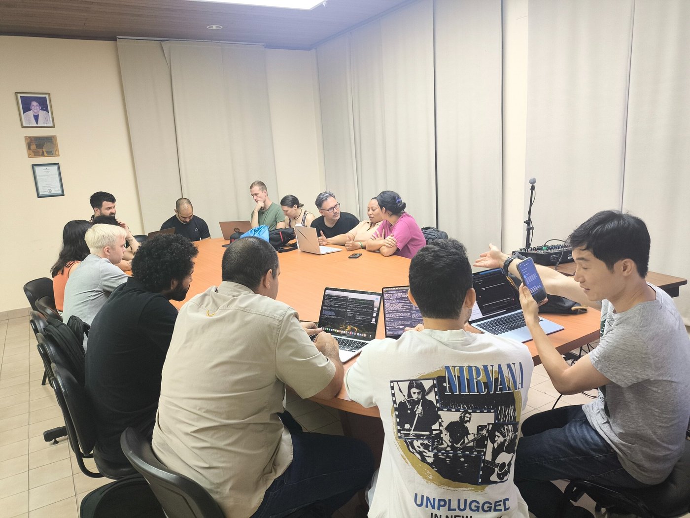(Photo) A group of people are gathered around a large wooden conference table in a meeting room, engaged in a discussion with several open laptops and one person showing something on a smartphone. Text: NIRVANA UNPLUGGED IN NEW YORK people, meeting, discussion | indoor | group around a conference table, laptops, smartphone | candid | Colors: #D2B48C, #FFFFFF, #000000, #E5E5E5 Note: The image is a real-life photograph capturing a group of people in an indoor meeting setting.