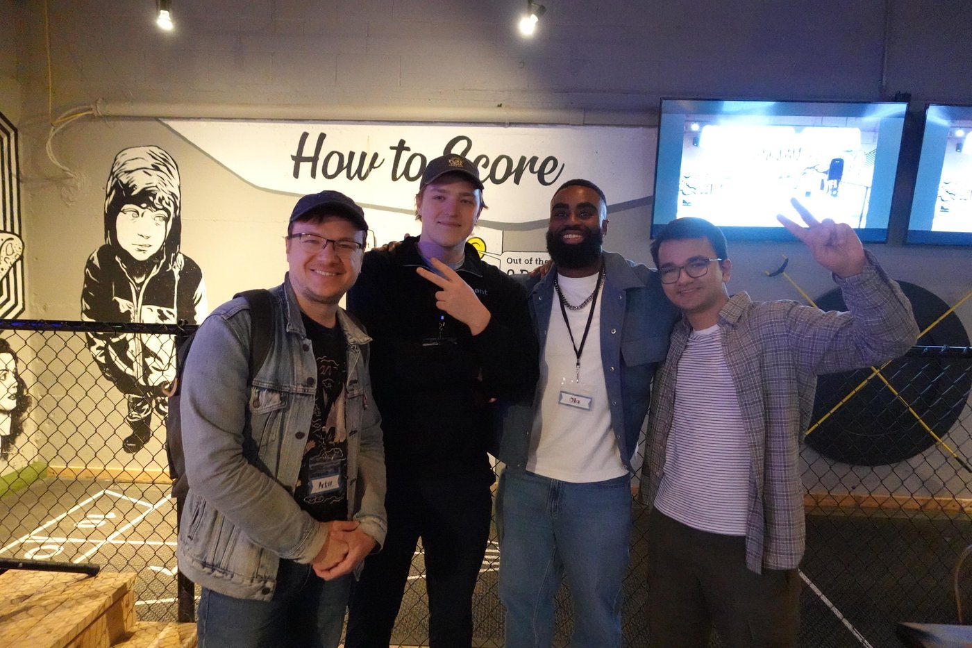 (Photo) A group photo of four men posing together indoors in front of a wall with a mural and the text 'How to Score'. Text: How to Score Out of the people | indoor | four men, mural background, chain-link fence, indoor lighting | group portrait | Colors: #1c1c1e, #8ea4c1, #e5e5e7, #4a4a4a Note: The image is a real-life photograph capturing a group of people posing together in an indoor setting.