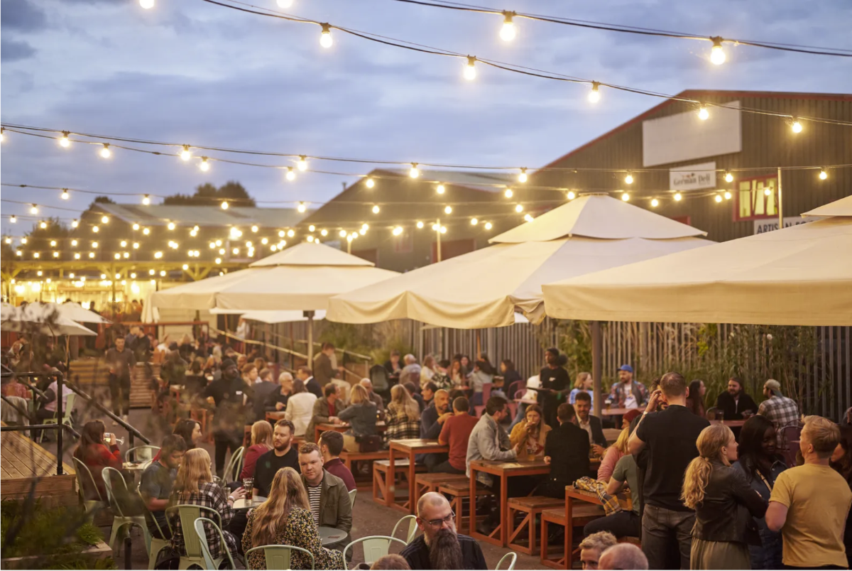 A photo of an outdoor dining area at dusk, filled with people seated at tables under string lights and umbrellas.