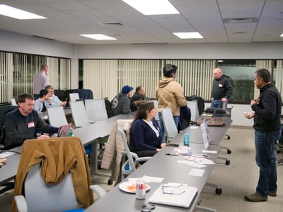 A group of people are gathered in an office or classroom setting, with some individuals sitting at long tables working on laptops and others standing and engaged in conversation.