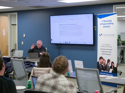 A man is giving a presentation in an office setting, with a large screen displaying text and a promotional banner for IT recruiting nearby.