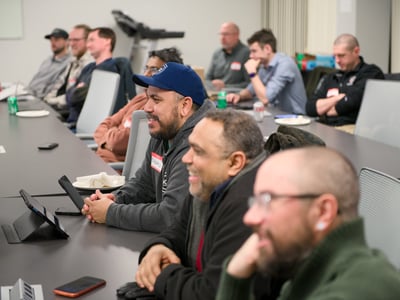 A group of men are seated at long tables in a conference room, attentively watching a presentation or speaker off-camera.