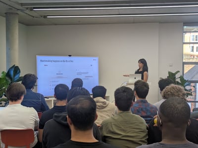 An indoor photograph showing a woman giving a presentation with a laptop and a large screen displaying software to an audience seated in front of her.