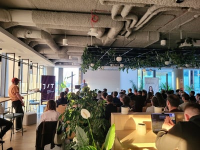 A large group of people are seated in a modern, industrial-style office space, attending a presentation given by a speaker at the front of the room.