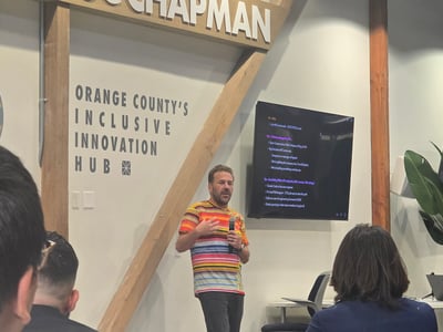 A man in a colorful striped shirt is giving a presentation in front of a screen and a wall labeled 'Orange County's Inclusive Innovation Hub' at Chapman University.
