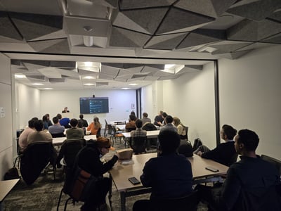 A group of people are seated in a modern conference room with unique geometric ceiling panels, attending a presentation displayed on a large screen.